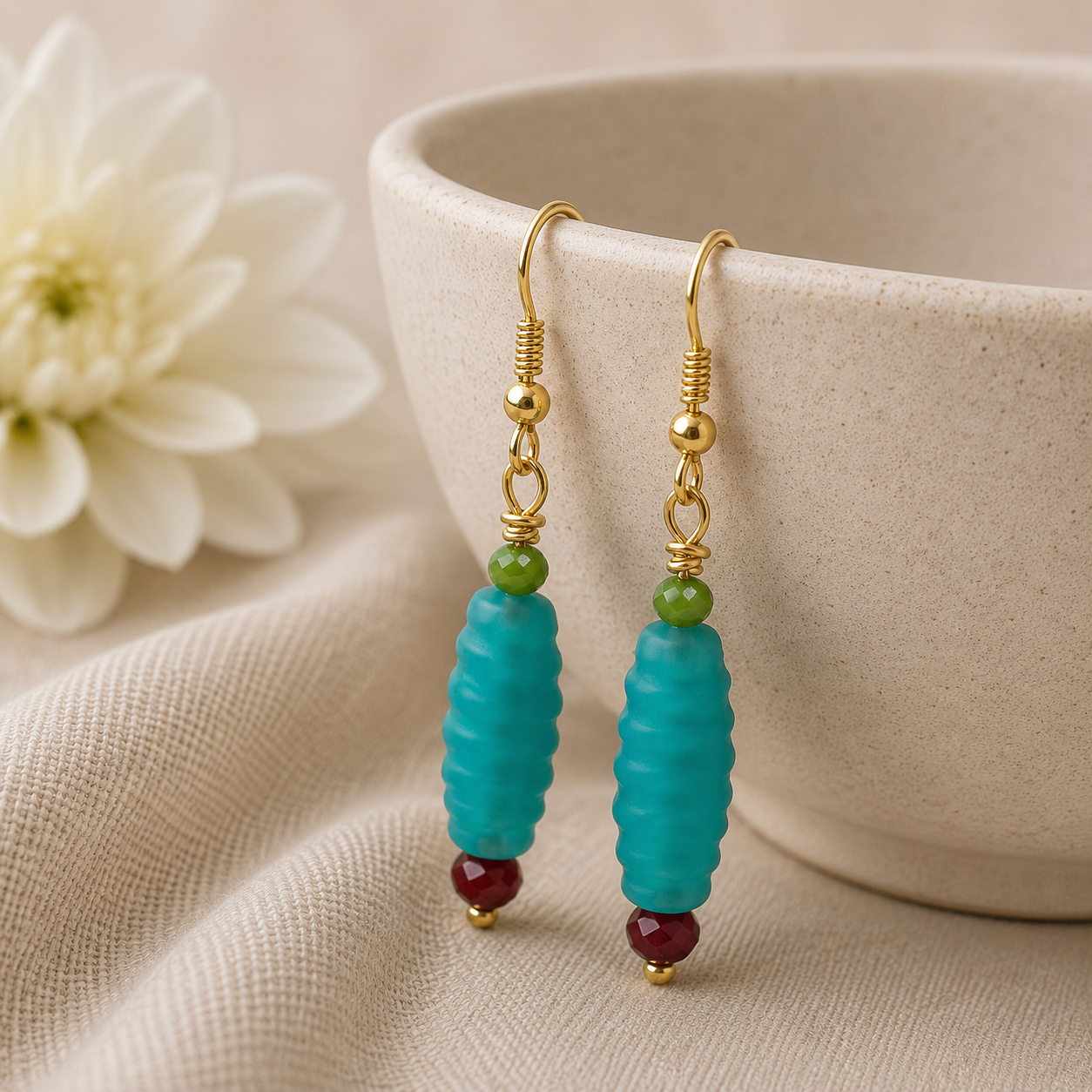 Turquoise beaded earrings with gold hooks on a beige surface with a white flower and bowl in the background.