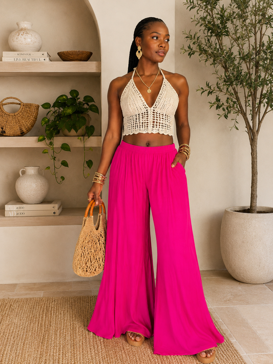 Woman in a white halter top and pink wide-leg pants holding a woven bag indoors.